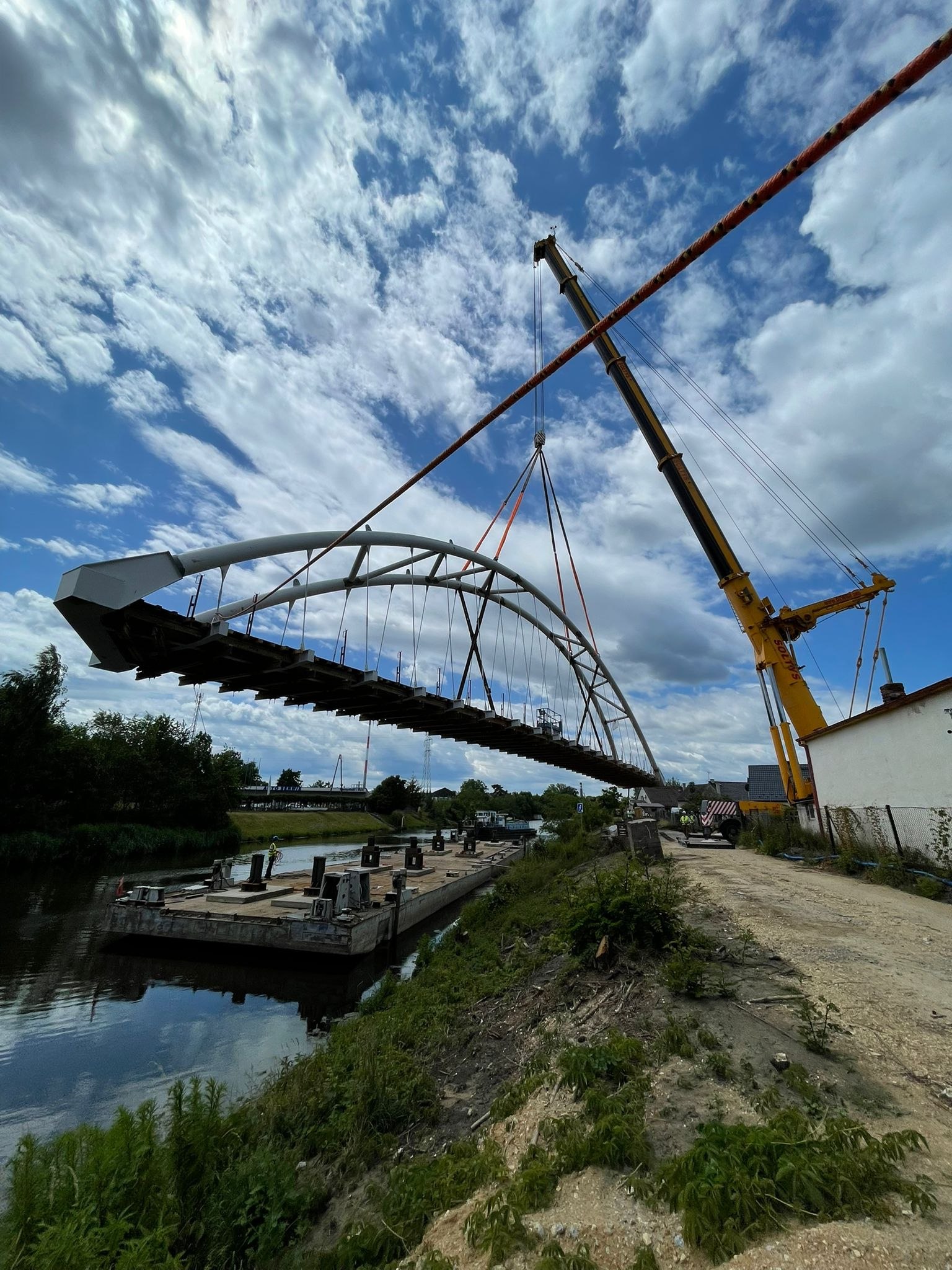 Dźwig Liebherr ustawia stalowe przęsło mostu łukowego nad rzeką, na potrzeby montażu na barkach.