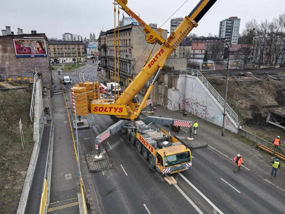 Żółty żuraw samojezdny Sołtys rozstawiony na środku miejskiej ulicy, pod wiaduktem kolejowym, w otoczeniu robotników w kaskach.