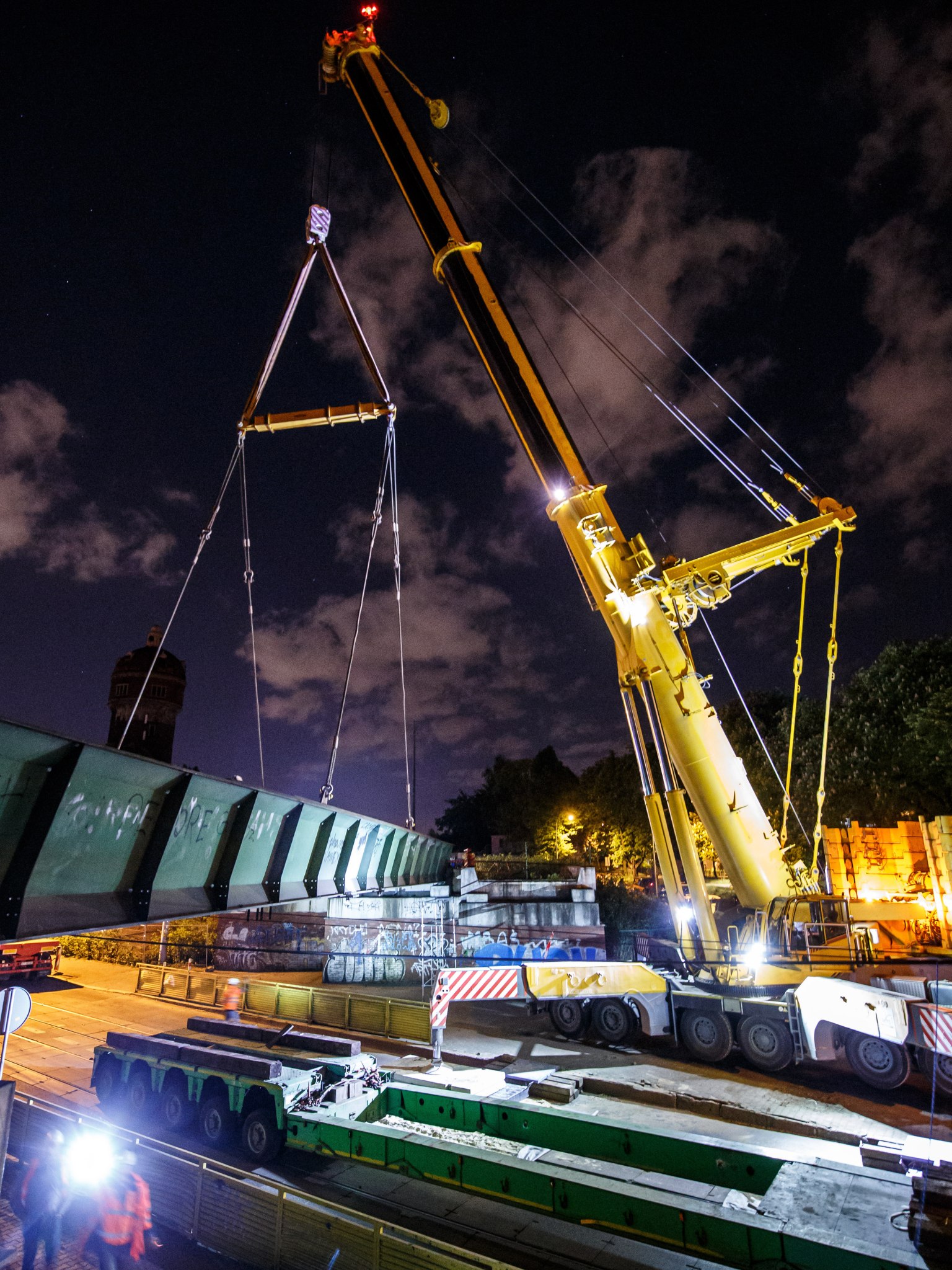 W nocy żuraw samojezdny podnosi stalowe przęsło mostowe nad ulicą, w świetle roboczych reflektorów i miejskich latarni.