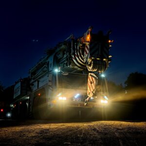 Mobilny dźwig Terex z włączonymi światłami stoi na nieoświetlonym placu budowy o zmierzchu, gotowy do pracy nocnej.