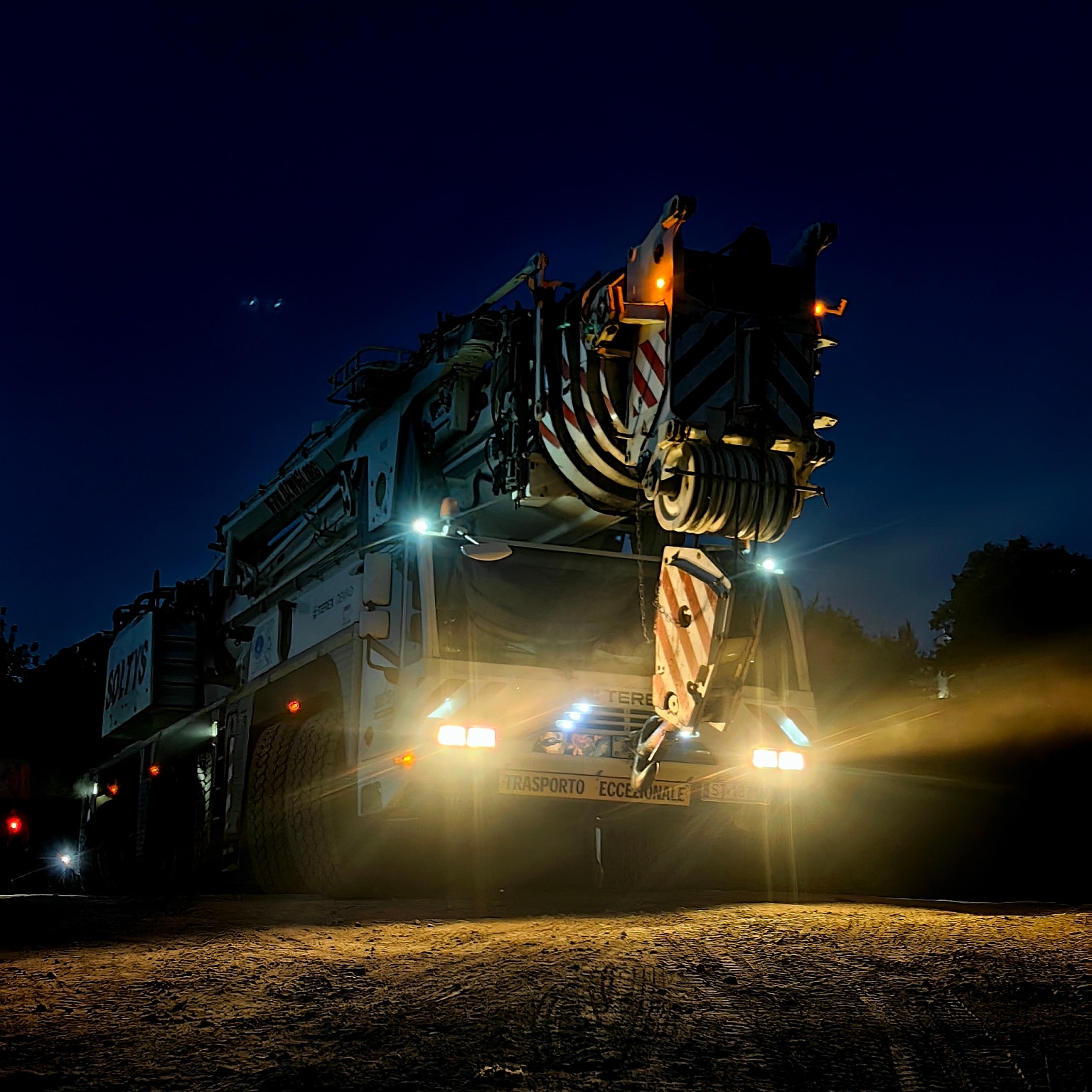 Mobilny dźwig Terex z włączonymi światłami stoi na nieoświetlonym placu budowy o zmierzchu, gotowy do pracy nocnej.