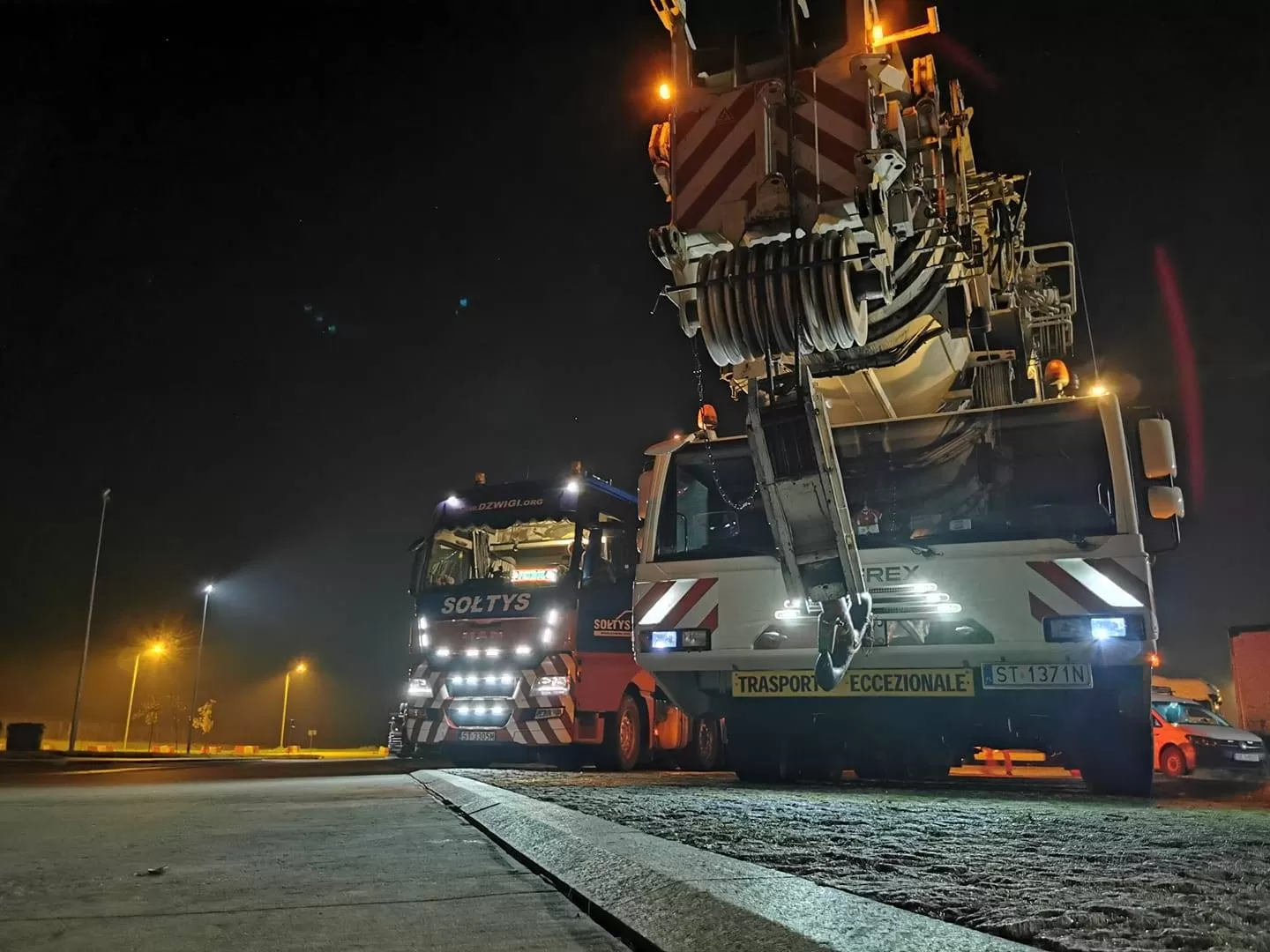 Ciężarówka transportowa do balastów i dźwig samochodowy samojezdnyh SOŁTYS przygotowane do transportu nocą na oświetlonym parkingu.